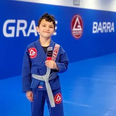 Un garçon en kimono bleu, ceinture grise, devant le logo Gracie Barra
